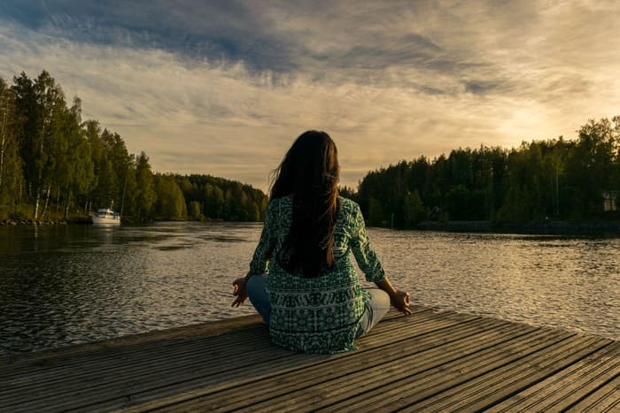 Femme méditant au bord d'un lac au coucher du soleil, illustrant le lâcher-prise, la relaxation et le bien-être grâce à l'hypnose et à la sophrologie