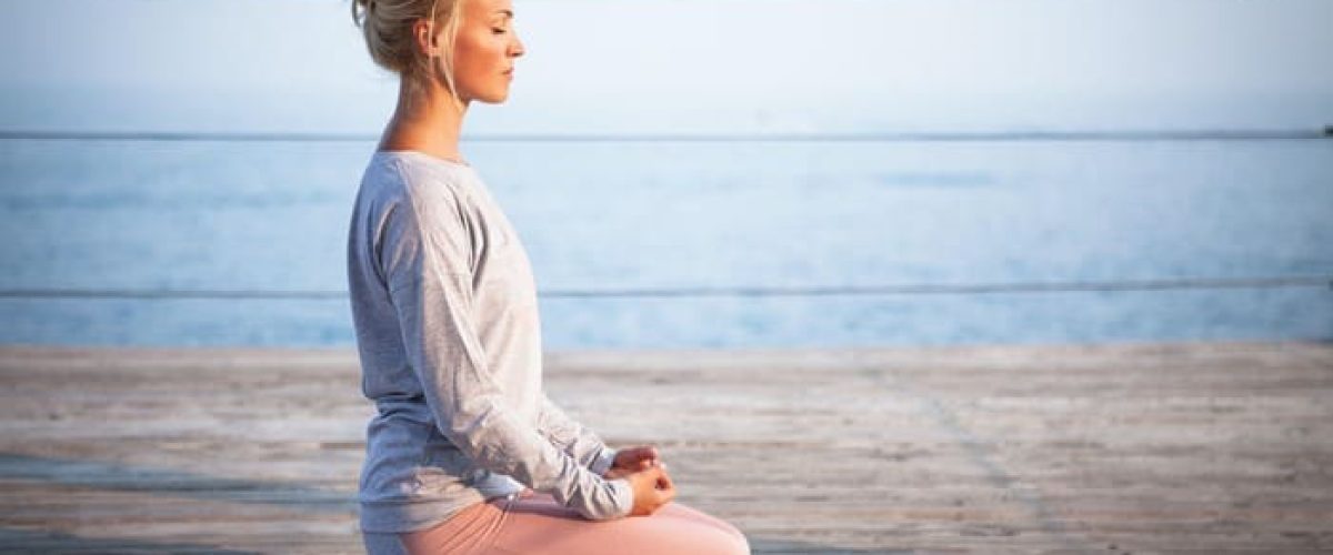 Femme pratiquant un exercice de respiration et de relaxation en bord de mer, illustrant une séance de sophrologie.